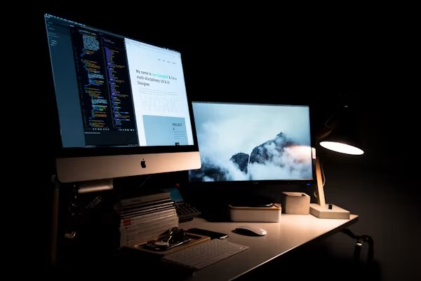 Abgedunkelter Schreibtisch bei Nacht: auf einem Bildschirm Quelltext neben der fertigen Website, daneben ein zweiter Monitor und eine Schreibtischlampe